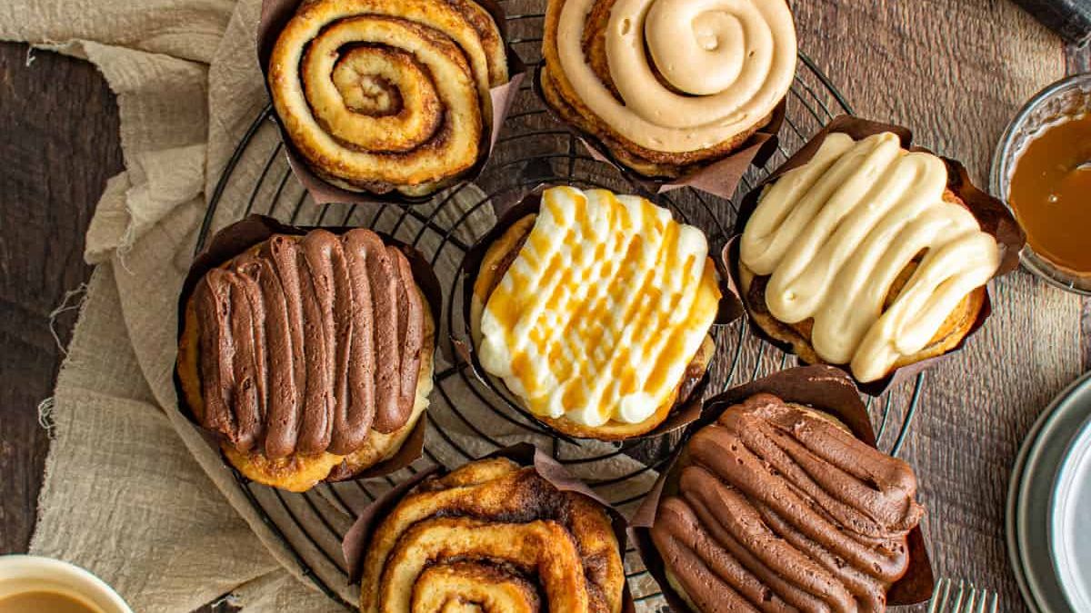 A close-up of a tray containing six cinnamon rolls. The rolls have different toppings, including chocolate, vanilla, and caramel icing, with some having a drizzle pattern. A cup of coffee and a jar of caramel sauce are partially visible in the background.