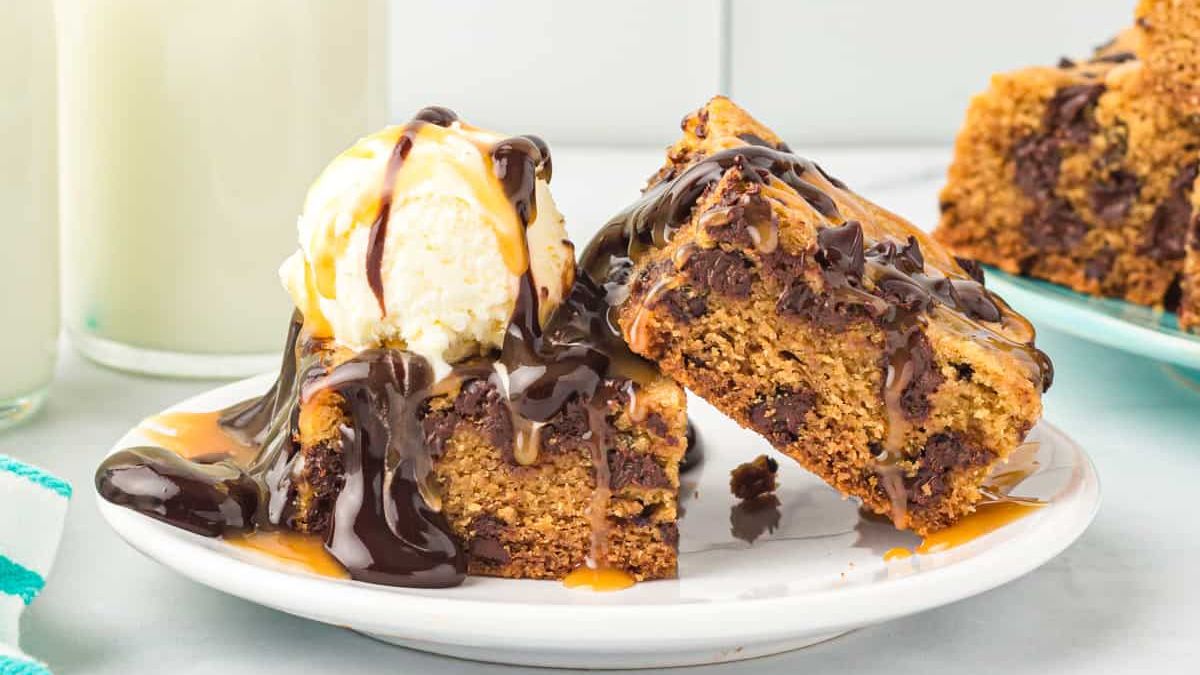 A dessert plate featuring two peanut butter chocolate chip cookie bars topped with a scoop of vanilla ice cream. The bars and ice cream are drizzled with chocolate and caramel sauce. A partly visible napkin and glass of milk are in the background.