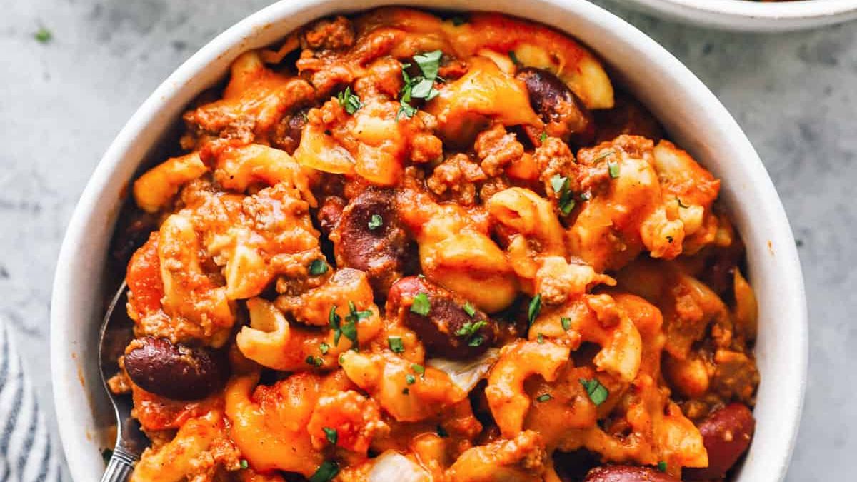 A bowl of cheesy pasta with ground meat, kidney beans, and chopped vegetables, garnished with herbs. The pasta appears to be mixed with a rich tomato-based sauce. A spoon is placed inside the bowl, ready to serve.