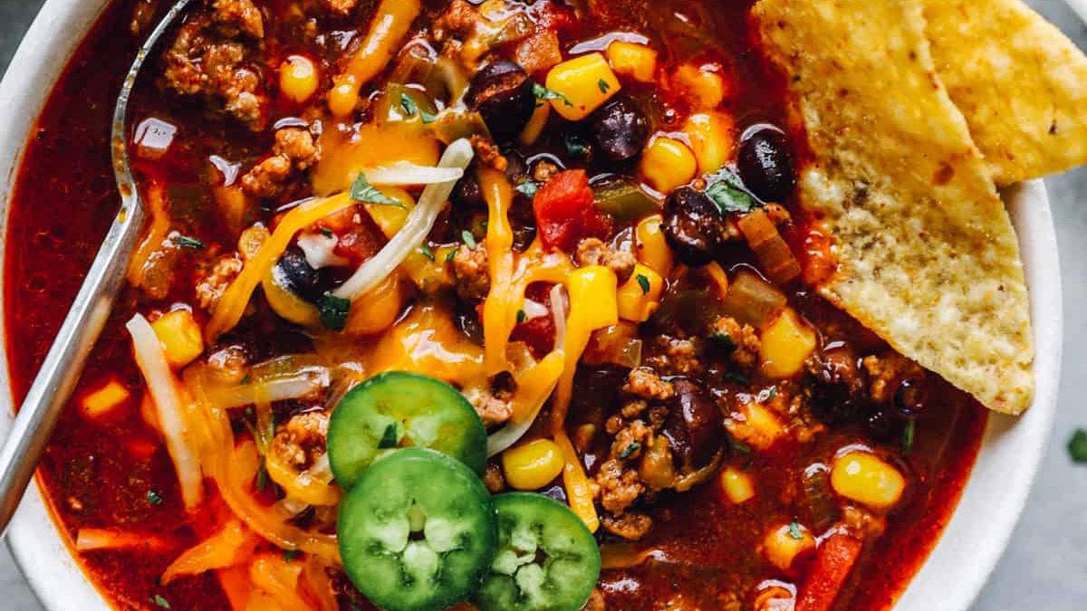 A bowl of hearty chili topped with sliced jalape&ntilde;os, shredded cheese, and chopped cilantro. The chili contains ground meat, black beans, corn, tomatoes, and diced vegetables. A metal spoon is in the bowl, and two tortilla chips are placed on the side.