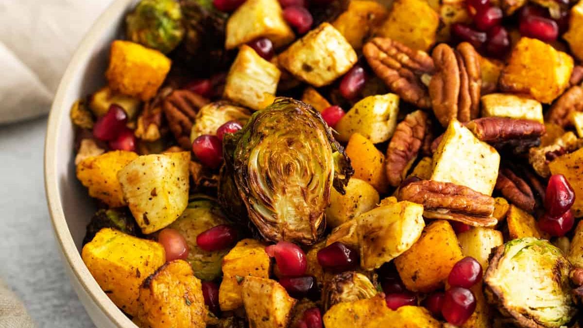 A close-up of a bowl filled with roasted vegetables, including Brussels sprouts, butternut squash, and root vegetables. Topped with pecans and pomegranate seeds for garnish. The colors are vibrant, featuring greens, yellows, and reds.