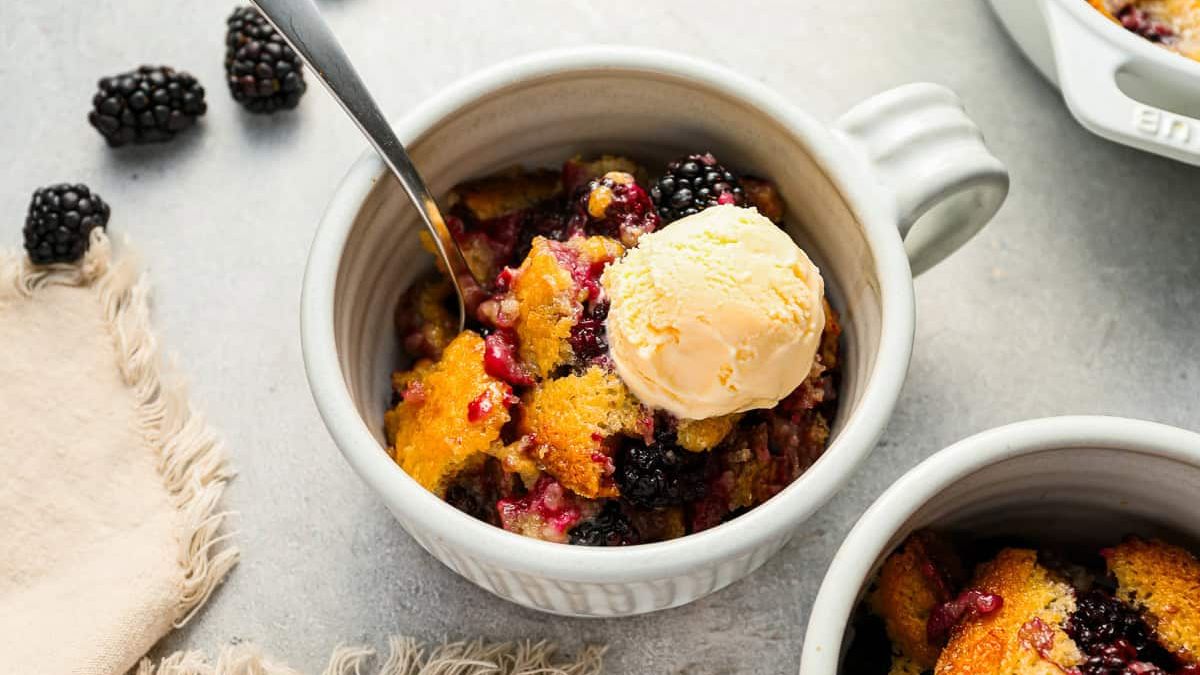 A mug filled with warm berry cobbler, topped with a scoop of vanilla ice cream. Fresh blackberries are scattered around, and a spoon is partially submerged in the cobbler. A beige cloth napkin is nearby on a light gray surface.