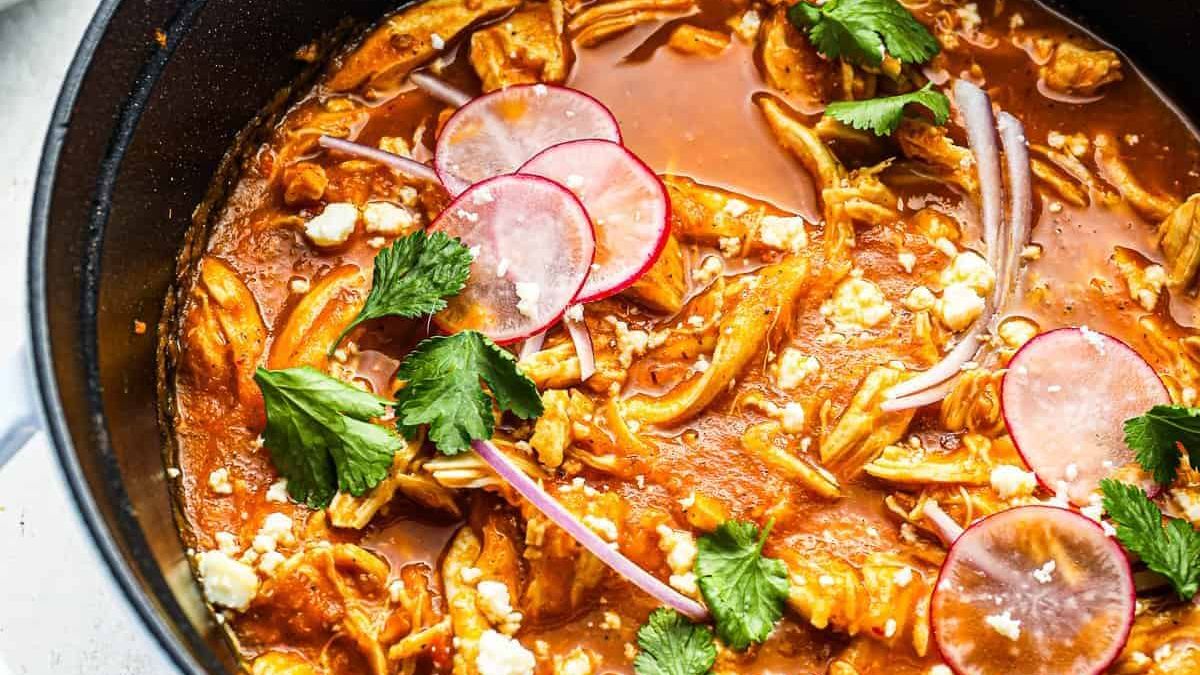 A close-up of a rich, flavorful soup in a black pot. The soup contains shredded chicken, topped with cilantro, crumbled cheese, thin radish slices, and thinly sliced red onions, all bathed in a vibrant red broth.
