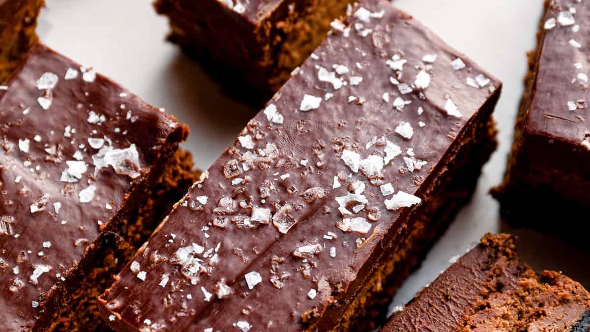 Close-up of several chocolate brownies topped with a glossy layer of chocolate icing and sprinkled with coarse sea salt. The brownies are cut into rectangular pieces and arranged on a flat surface.