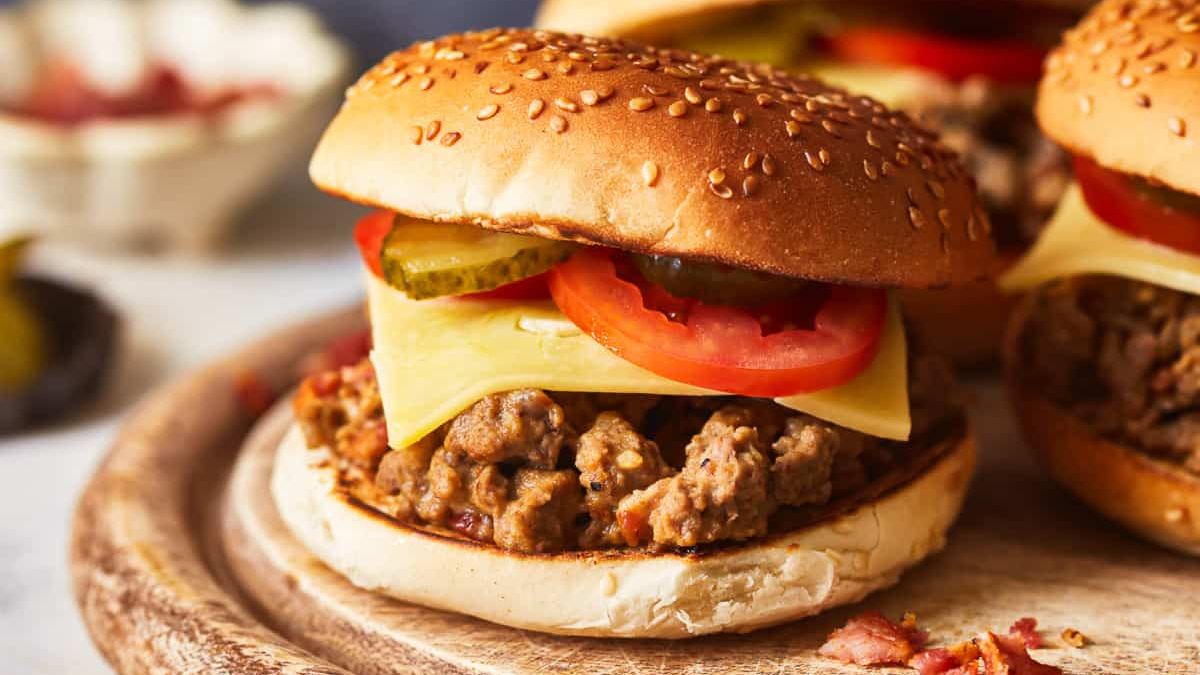 A close-up of a juicy burger on a sesame seed bun sits on a wooden board. The burger is topped with slices of tomato, pickles, and melted cheese. In the background, another burger and some blurred vegetables can be seen.
