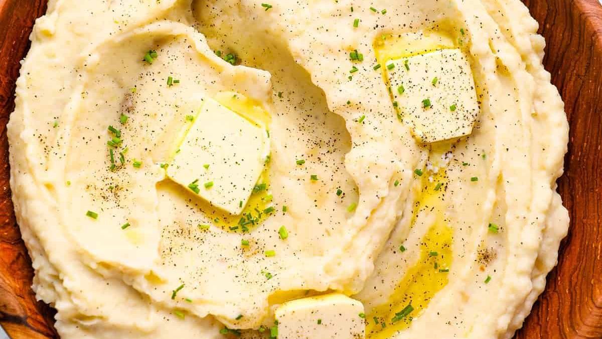 A wooden bowl filled with creamy mashed potatoes, topped with three small melted butter pats, black pepper, and chopped green herbs. The mashed potatoes have a smooth and fluffy texture, with visible swirls from spoon mixing.