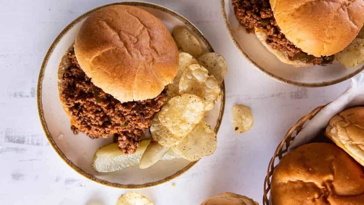 A plate with a sloppy joe sandwich and a pickle spear placed beside a pile of potato chips. Another plate with a similar arrangement is partially visible at the top right, and part of a bread basket is seen at the bottom right.