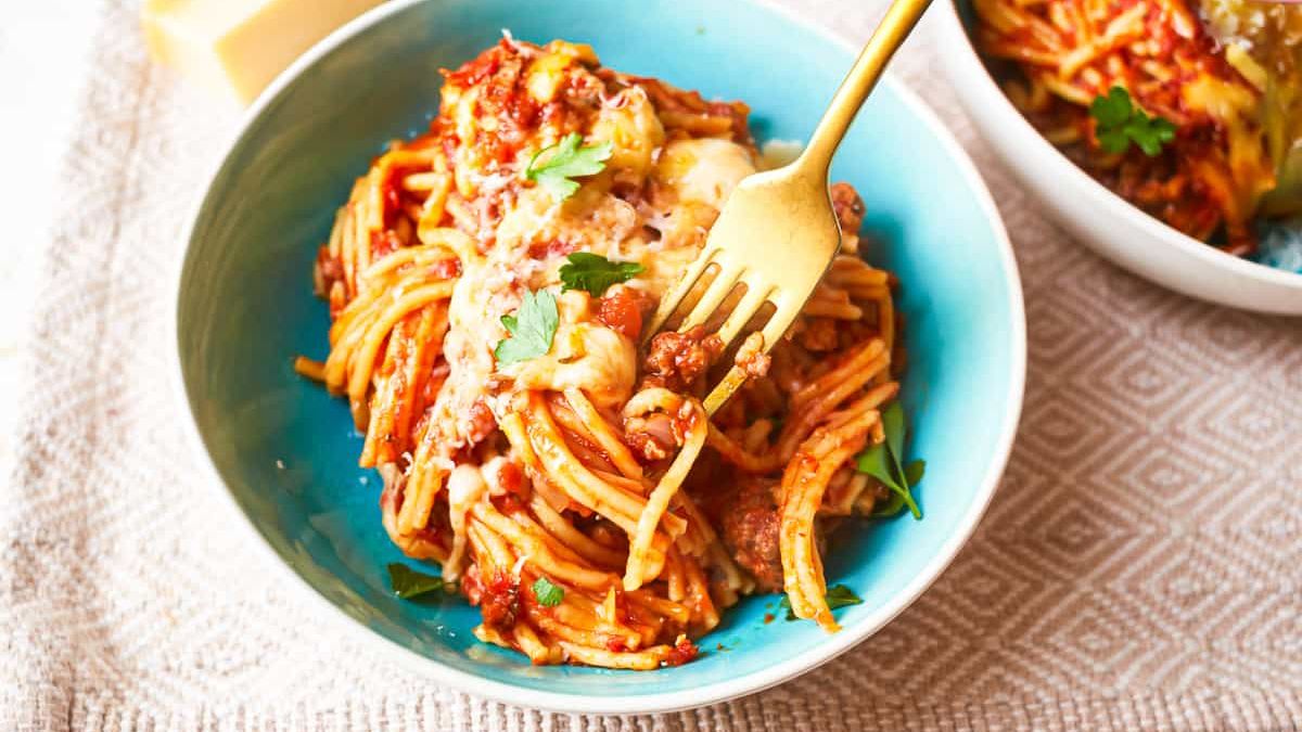 A blue bowl filled with spaghetti topped with tomato sauce, grated cheese, and garnished with fresh parsley. A gold fork is twirling a portion of the spaghetti. The bowl is placed on a light-colored textured mat. Another bowl of spaghetti is partially visible in the background.