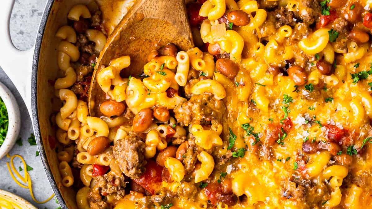 A skillet filled with cheesy macaroni mixed with ground beef, beans, and diced tomatoes. A wooden spoon is stirring the mixture, which is garnished with fresh parsley.