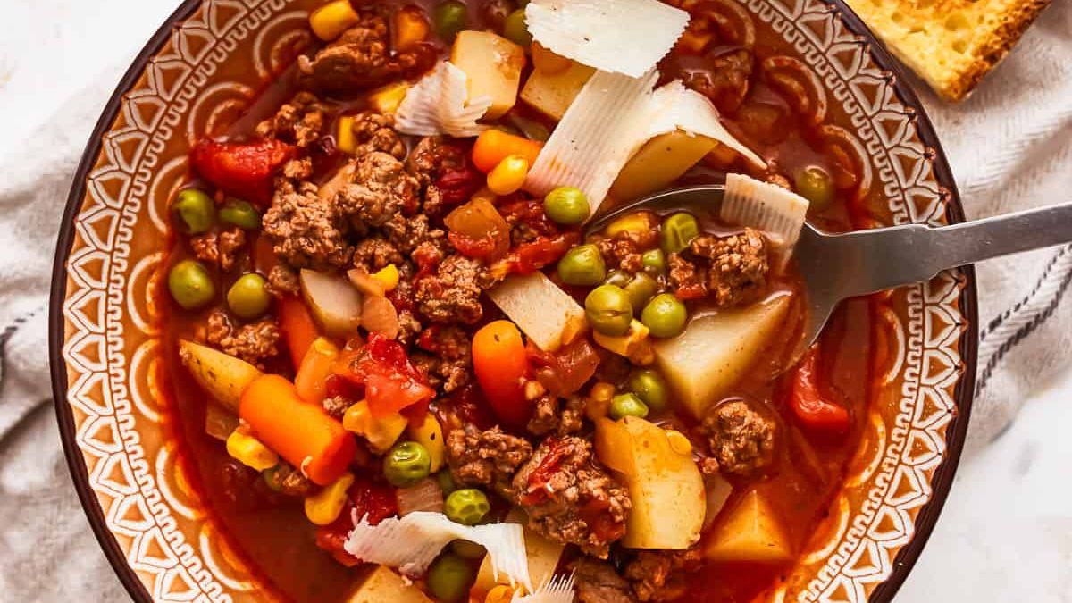 A hearty bowl of vegetable beef soup filled with ground beef, potatoes, carrots, peas, corn, and tomatoes, topped with thinly shaved cheese slices. A piece of cornbread sits partially out of the frame next to the bowl, and a spoon is resting in the soup.