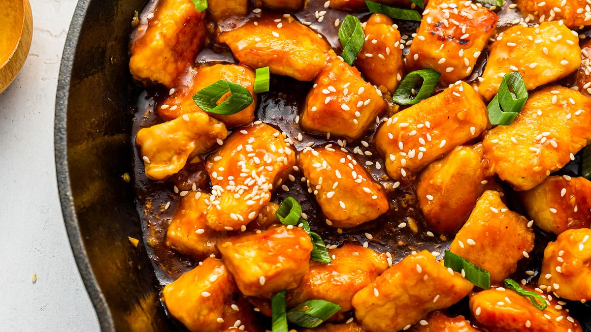 A close-up of sizzling honey garlic chicken in a skillet. The chicken pieces are glazed with a golden-brown sauce, garnished with sesame seeds and chopped green onions.