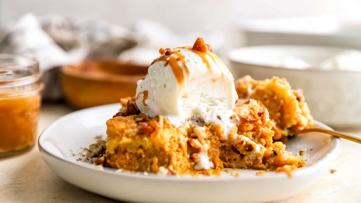 A delicious dessert consisting of a pumpkin crunch cake topped with a scoop of vanilla ice cream and a drizzle of caramel sauce. The dish is served on a white plate with a blurred background that includes a jar of caramel and a bowl.