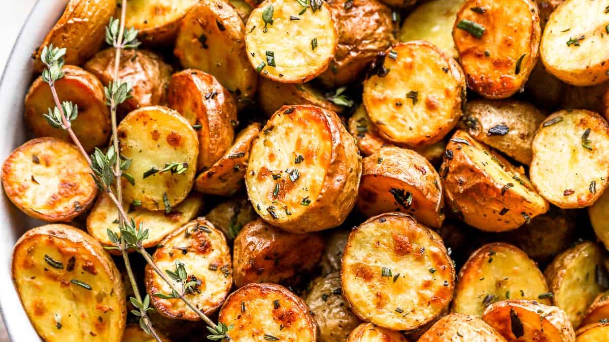 A close-up of a dish of golden, roasted baby potatoes garnished with fresh herbs such as rosemary and thyme. The potatoes are crispy and slightly charred, suggesting they have been roasted to perfection.
