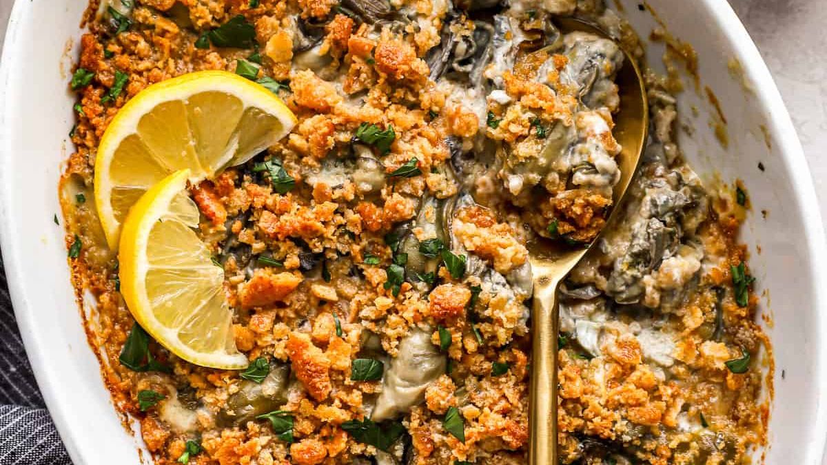 A creamy baked dish with oysters, topped with a golden breadcrumb crust. Garnished with parsley and lemon slices on the side, a gold spoon rests in the dish. Served in an oval white baking dish.