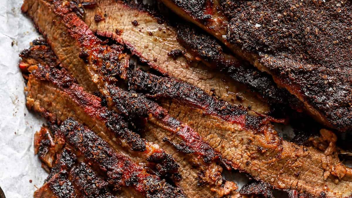 Close-up of juicy, sliced smoked beef brisket on parchment paper. The slices are charred and seasoned on the outside, revealing a tender, well-cooked interior with a deep, rich color.