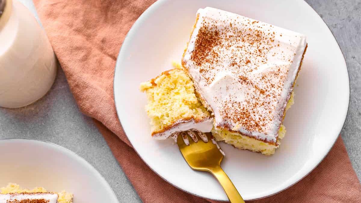 A slice of frosted vanilla cake topped with cocoa powder on a white plate, with a fork holding a piece of the cake. The plate is placed on a peach napkin with a glass of milk beside it, all set on a light-colored surface.
